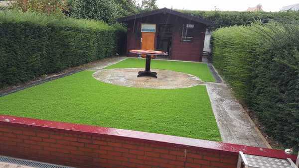 Lage rode bakstenen muur met daarachter een tuin met kunstgras, met in het midden een tafel en achterin de tuin een houten tuinhuis