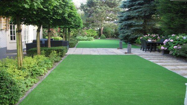 Tuin met aan de linkerkant bomen, in het midden een strook kunstgras en aan de rechterkant betegeling met een tafel en stoelen